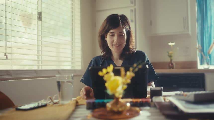 A female medical professional in scrubs smiles while studying on a tablet in her bright kitchen at home in slow motion