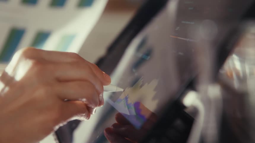A business professional analyzes financial data graphs on a digital tablet using a stylus in a naturally lit office