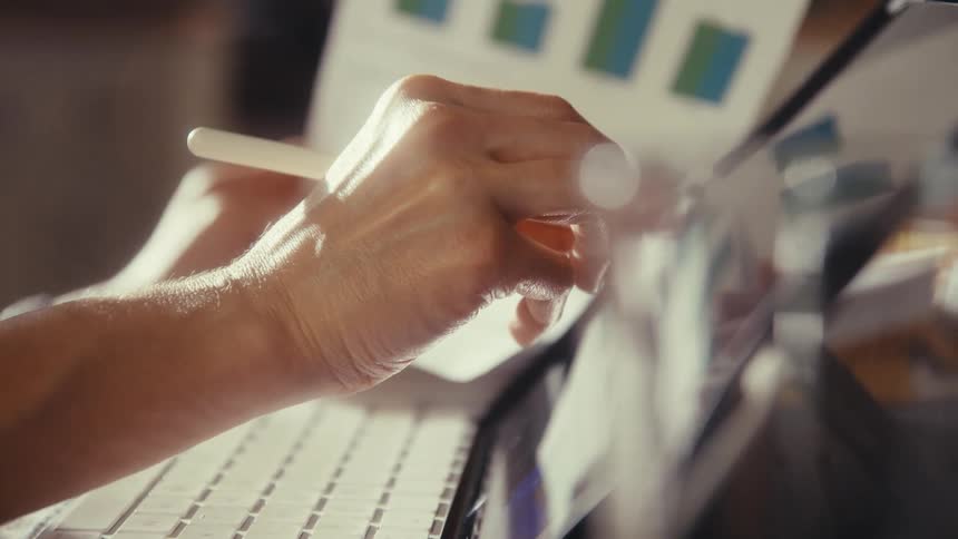 A person's hands use a stylus on a tablet screen while referencing a printed bar chart in a warm, naturally lit office setting