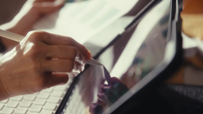 A detailed close-up shows a hand using a digital pencil on a tablet with a keyboard, working in warm sunlight