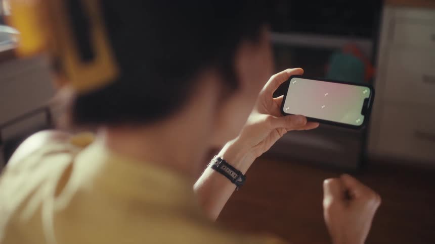 Over-the-shoulder view of a woman in a yellow top holding a smartphone horizontally with a green screen in her sunlit kitchen