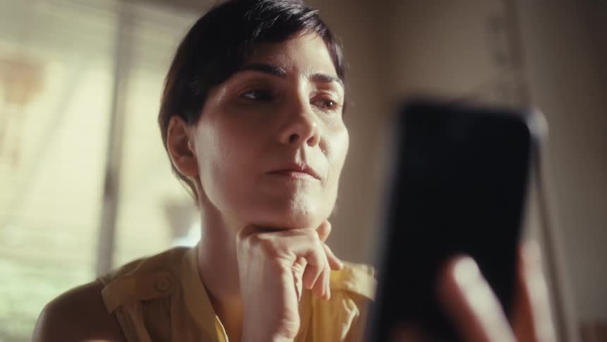 A close-up of a thoughtful woman scrolling on her smartphone, contemplating what she sees in a sunlit room