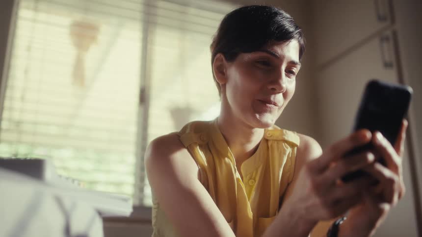 A joyful woman with short dark hair laughs while using her smartphone in a sunlit room