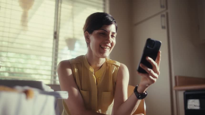 A cheerful woman in a yellow shirt enjoys watching content on her smartphone, smiling and laughing in a sunlit kitchen