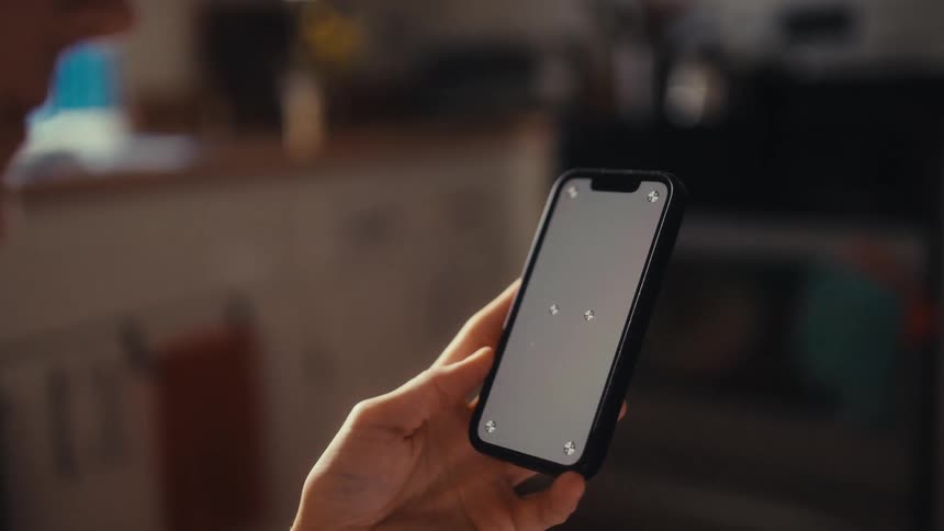 An over-the-shoulder close-up of a person holding a smartphone with a blank screen and tracking markers in a sunlit room