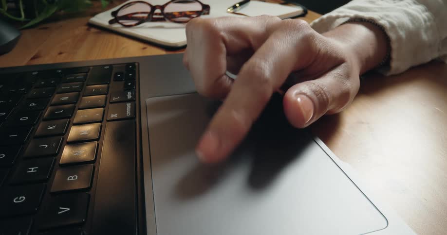 A close-up cinemagraph of a woman's finger scrolling on a laptop trackpad at her wooden desk