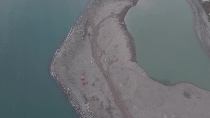 An aerial top-down view pulls back to reveal heavy construction equipment building a breakwater between the sea and a harbor