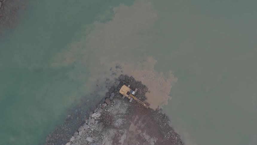 An aerial top-down view shows an excavator moving rocks and dirt on the edge of a murky body of water, creating clouds of sediment
