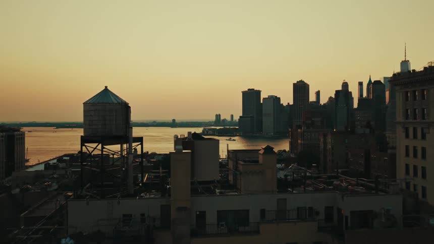 An aerial view flies over Brooklyn rooftops, revealing the iconic Manhattan skyline across the water at sunset