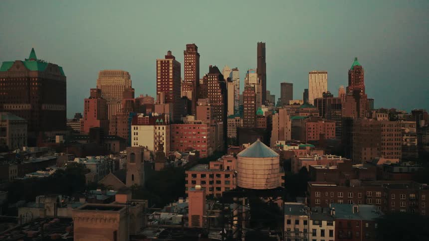 A slow aerial tracking shot reveals the Brooklyn skyline with historic architecture and a rooftop water tower at dusk