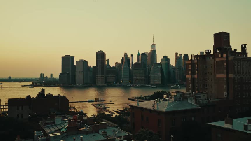 An aerial view of the Manhattan skyline at golden hour, pulling back to reveal Brooklyn rooftops and the East River
