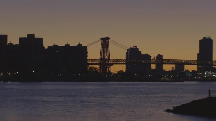 A zoom-out shot reveals the Williamsburg Bridge and New York City skyline silhouetted against a beautiful sunset sky over the East River