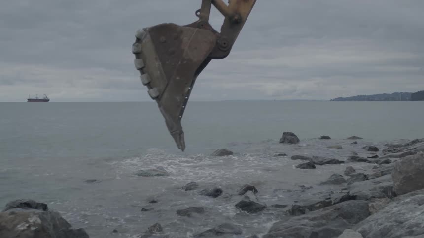 An excavator works on a rocky shoreline, dropping stones into the sea on a bleak, overcast day