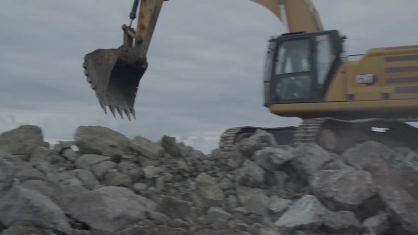 In slow motion, a powerful excavator lifts and moves a giant boulder at a construction site on a cloudy day