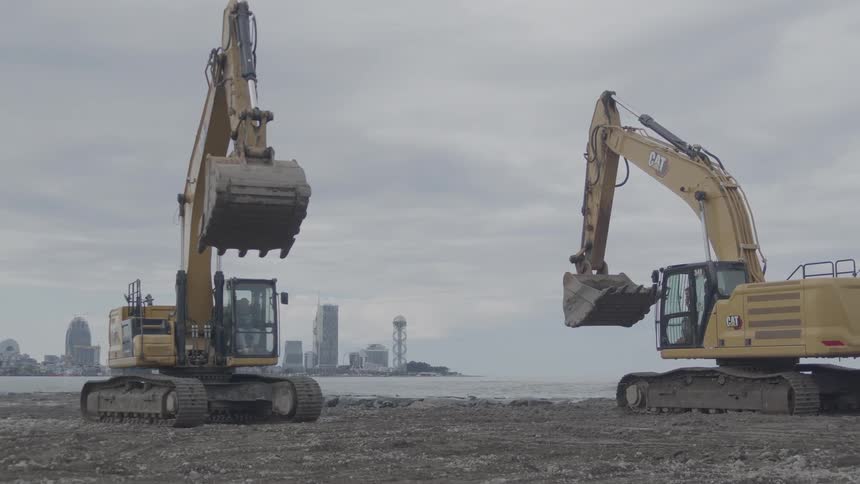 Two large yellow excavators sit on a coastal construction site with a modern city skyline across the water on an overcast day