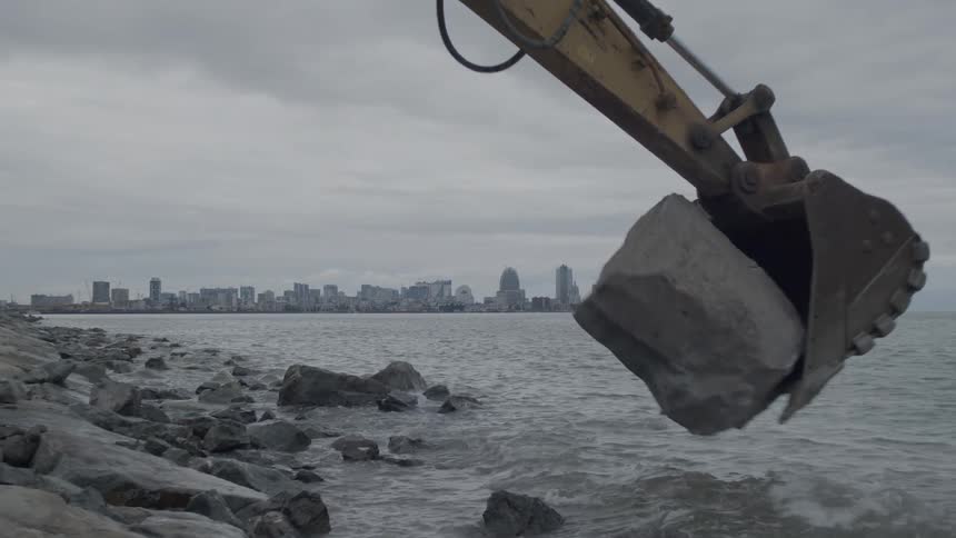 Slow motion shot of an excavator bucket dropping a large rock into the water to build a breakwater, with a city skyline in the background