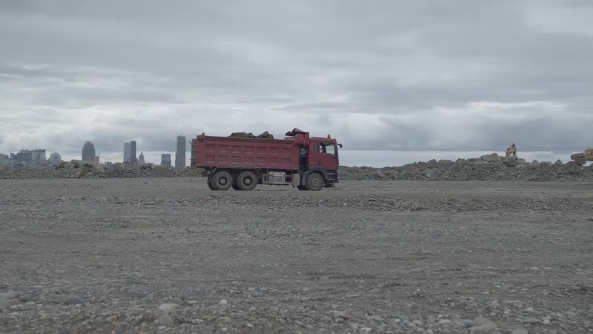 A large red dump truck reverses across a barren landscape with a modern city skyline in the distance