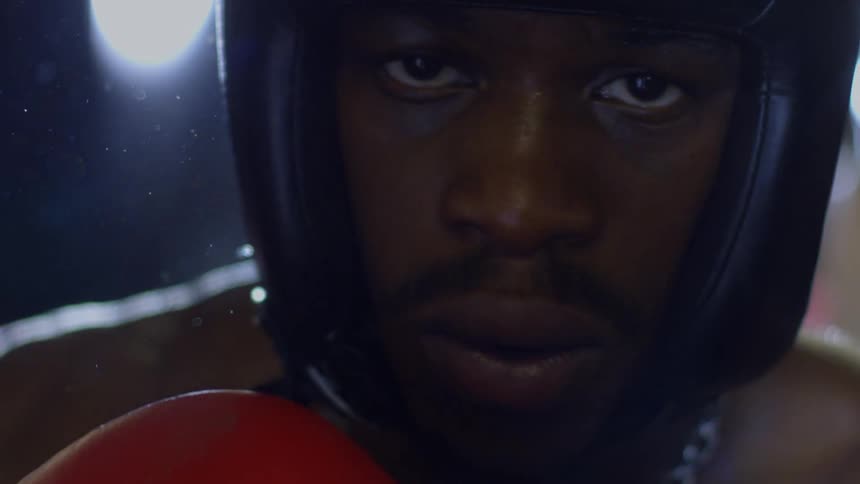 An intense, dramatic slow-motion close-up reveals a focused boxer's face in a helmet and gloves