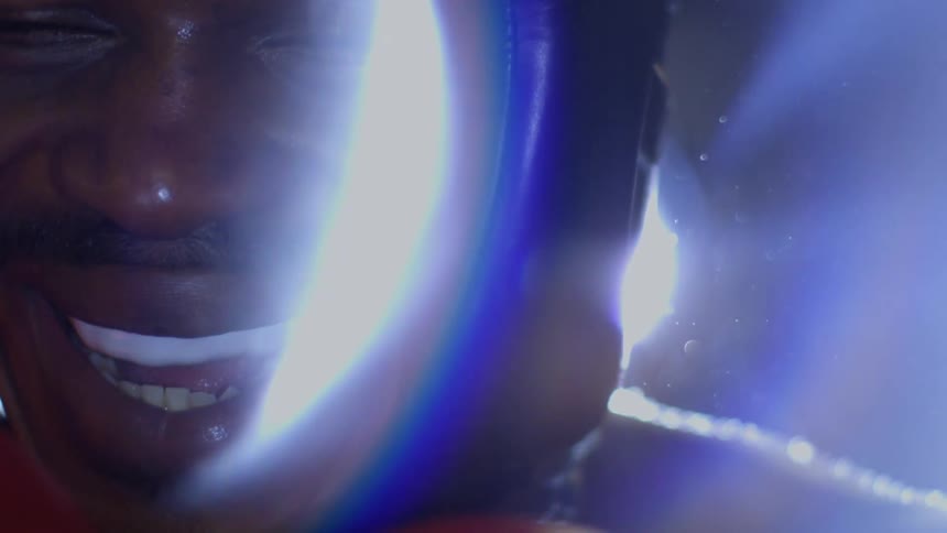 An extreme close-up of a boxer wearing a mouthguard and headgear as he breaks into a slow, confident smile