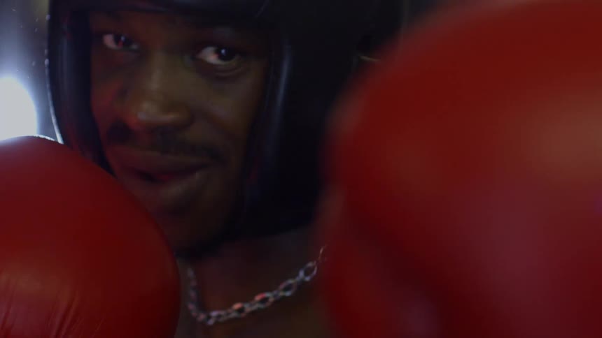 Dramatic close-up of a boxer in red gloves and headgear looking intensely at the camera with a slight smile