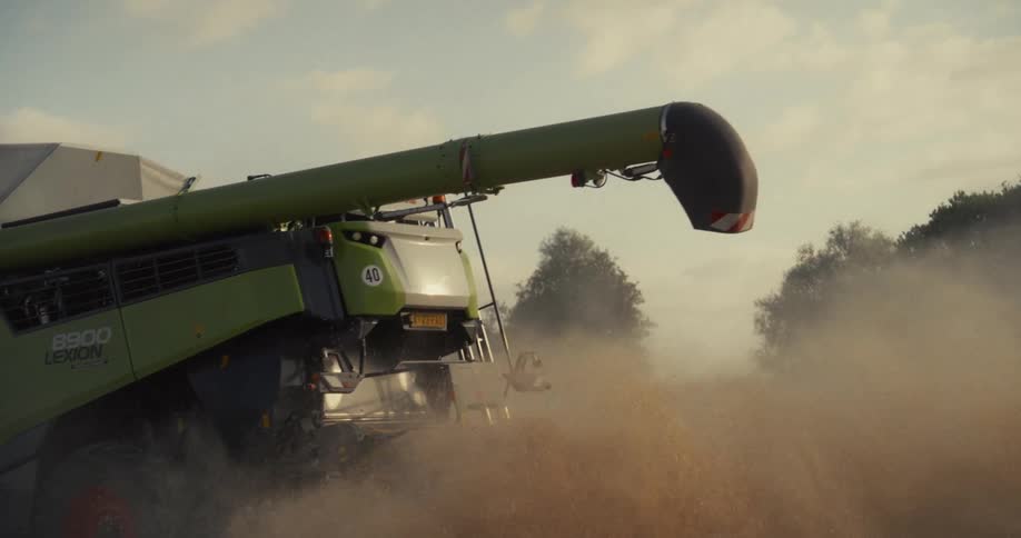 A powerful combine harvester works a field in slow motion, expelling a thick cloud of dust and chaff under the evening sun