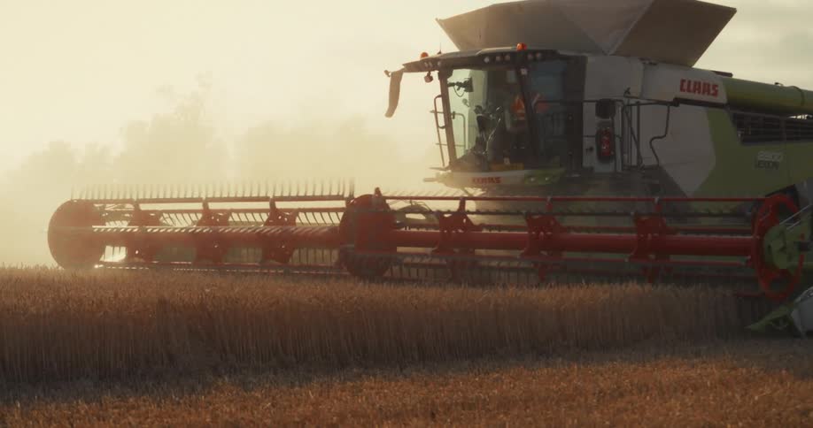A combine harvester moves through a golden field at sunset, kicking up dust in slow motion as it reaps the grain