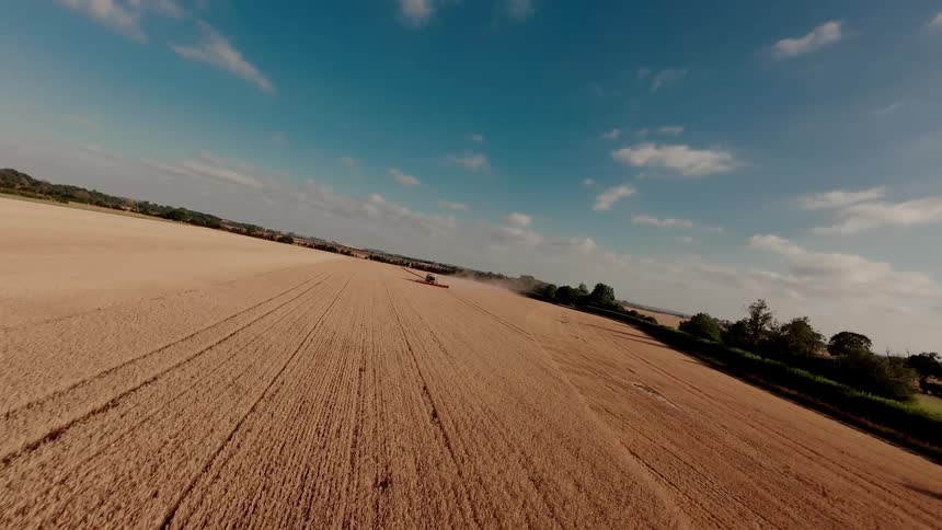 A tilted aerial perspective captures a combine harvester diligently working its way through a vast golden wheat field under a brilliant blue sky