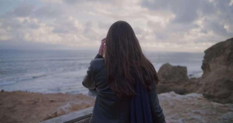 A pensive woman with long hair watches the ocean waves crash against the rocky shoreline from a cliffside viewpoint at dusk