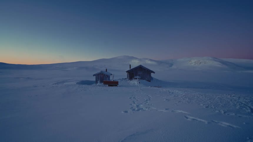 Two remote wooden cabins sit peacefully in a vast, snow-covered landscape during the tranquil blue hour at dusk