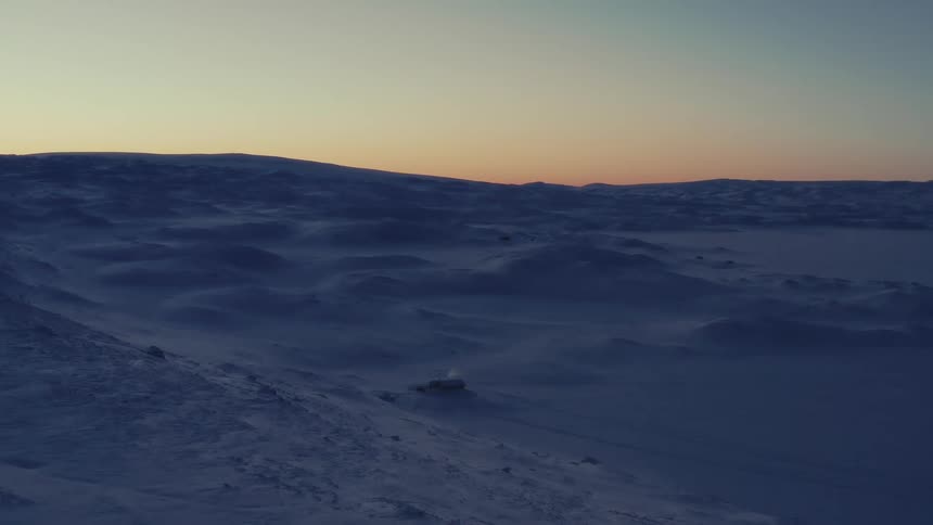 An isolated cabin emits smoke in a vast, snowy, and windswept arctic landscape during a cold sunset