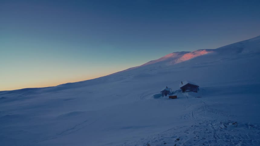 An aerial shot pulls back to reveal remote cabins nestled in a vast, snow-covered mountain landscape during a serene sunrise