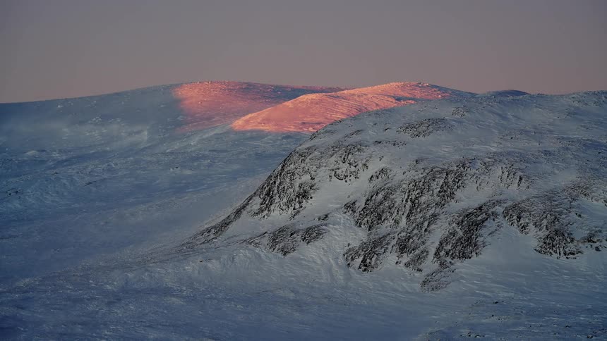 The peaks of a vast, snow-covered mountain range glow with a beautiful pink alpenglow at sunrise