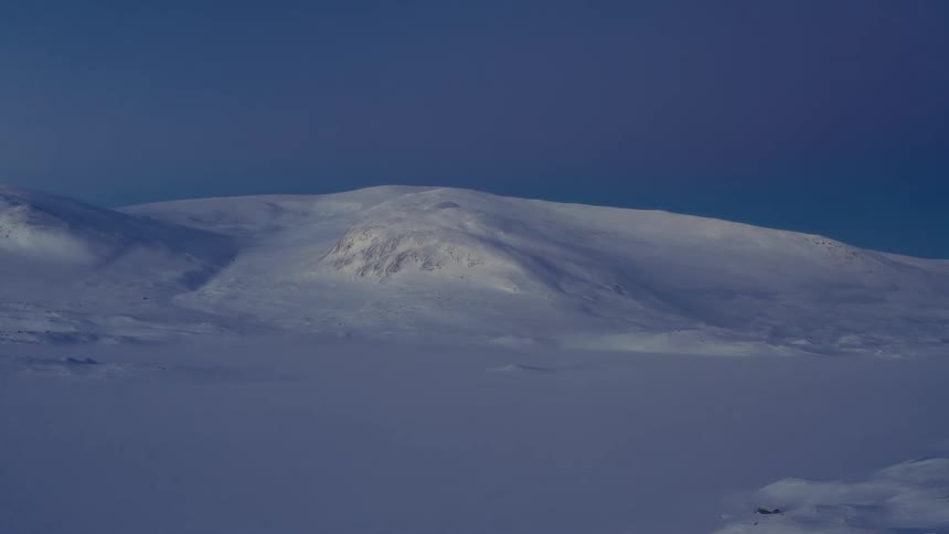 A slow zoom out reveals a vast, desolate landscape of snow-covered mountains as strong winds create a ground blizzard