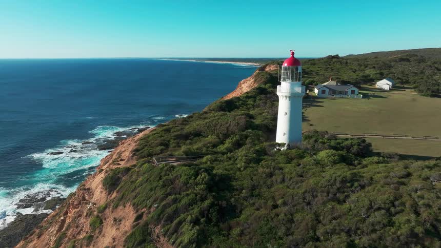 A scenic aerial view flies towards a majestic white lighthouse standing on a verdant cliff overlooking the vast blue ocean