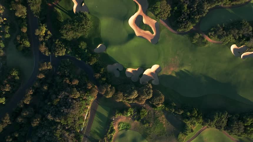 A stunning top-down aerial view reveals a lush green golf course with intricate sand traps bathed in the warm light of sunrise