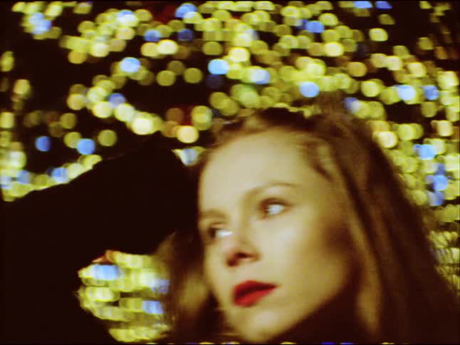 A glamorous blonde woman with red lipstick poses against a backdrop of twinkling holiday lights in a vintage 8mm film style