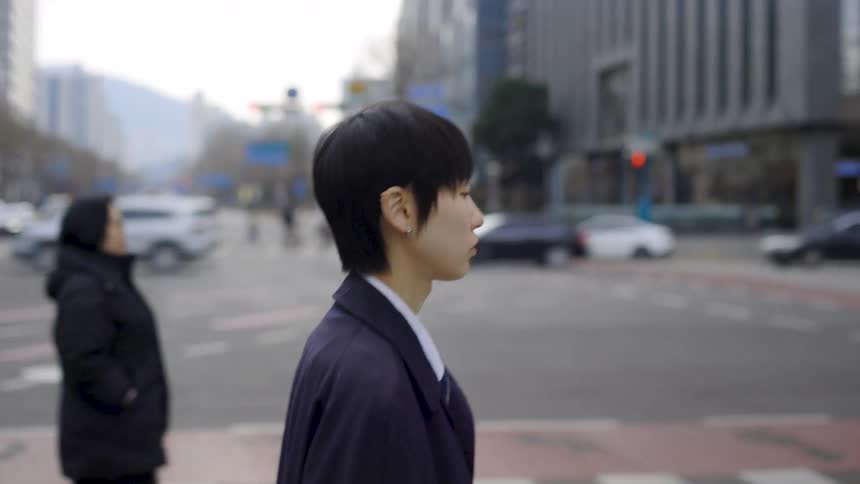 A young East Asian person with short hair walks across a busy city crosswalk in slow motion
