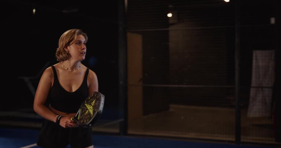 A female athlete in black sportswear serves the ball with focused determination on an indoor padel court in slow motion