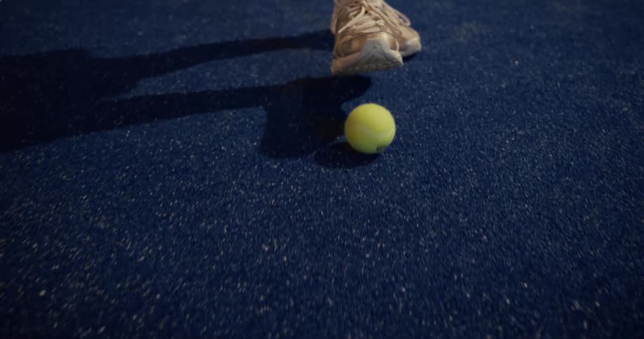 A close-up shot of a yellow tennis ball rolling across a blue court before being stopped by a player's shoe