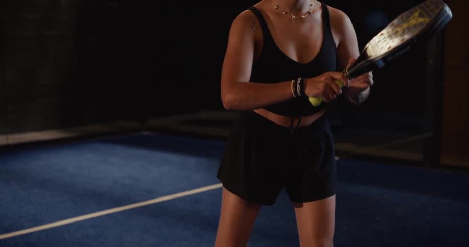 A female padel player in a black athletic outfit warms up by bouncing a ball on her racket in slow motion on an indoor court