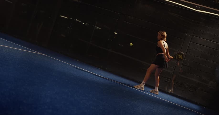 A female athlete serves the ball with precision during a padel tennis match, captured in dramatic slow motion on a blue court
