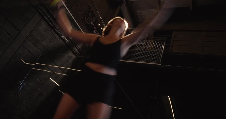 A woman serves a padel ball with motion blur on a dark indoor court, captured from a dramatic low angle