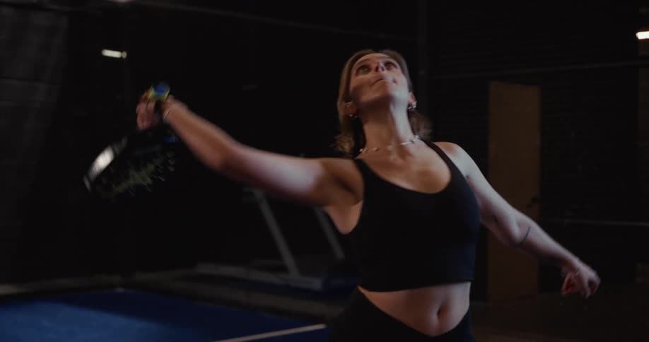 A focused female athlete serves a tennis ball with her racket in slow motion on a dark indoor court