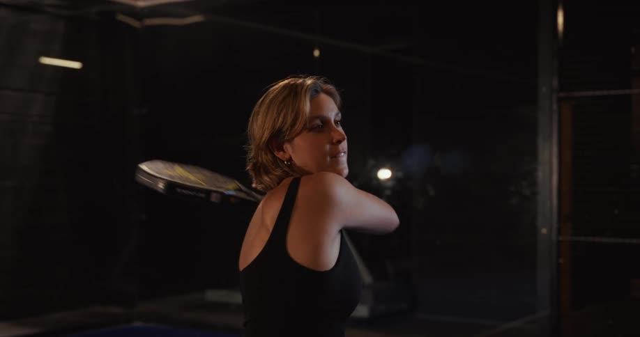 Slow motion shot of a woman serving a ball on a dark, indoor padel court