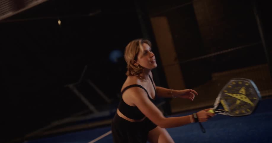 A determined female athlete in black sportswear plays padel tennis, swinging her racket at the ball in slow motion on a dark court