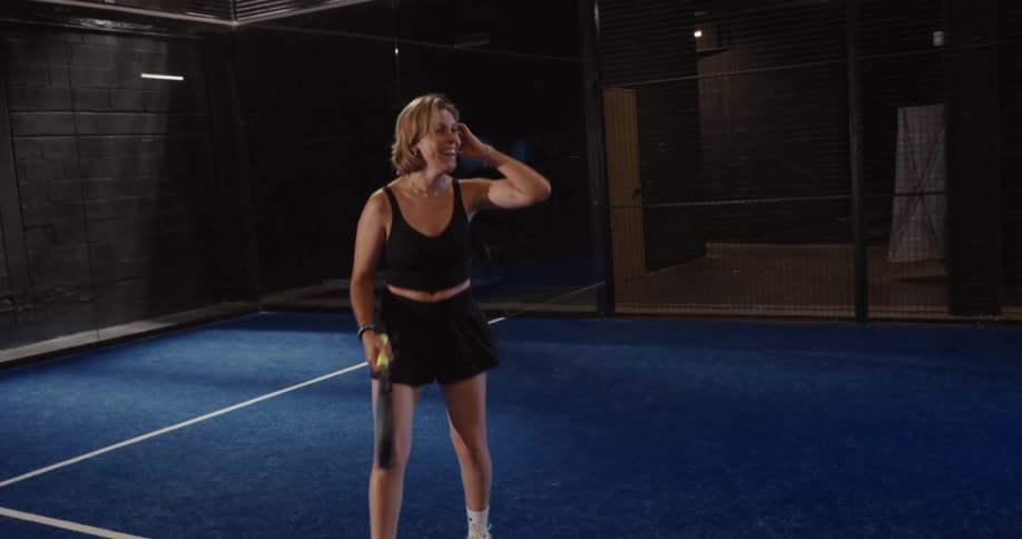 A smiling woman in sportswear hits a padel ball with her racket and laughs joyfully in slow motion