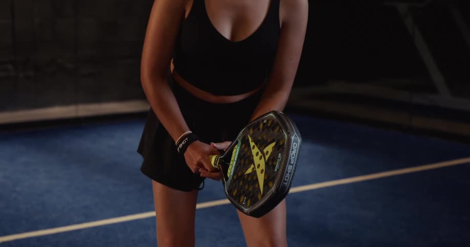A female athlete in black sportswear holds a padel racket in a ready stance on a blue indoor court