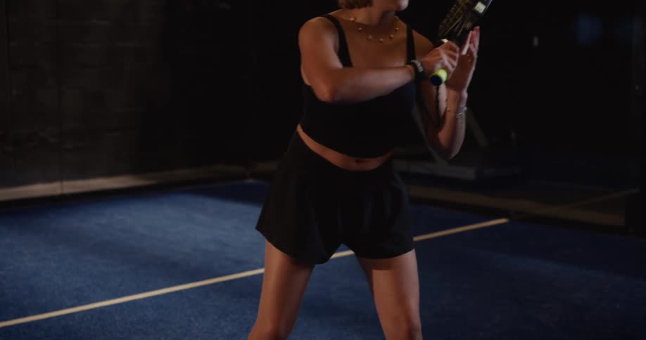 A female paddle tennis player warms up by bouncing the ball on her racket in slow motion on a dark indoor court