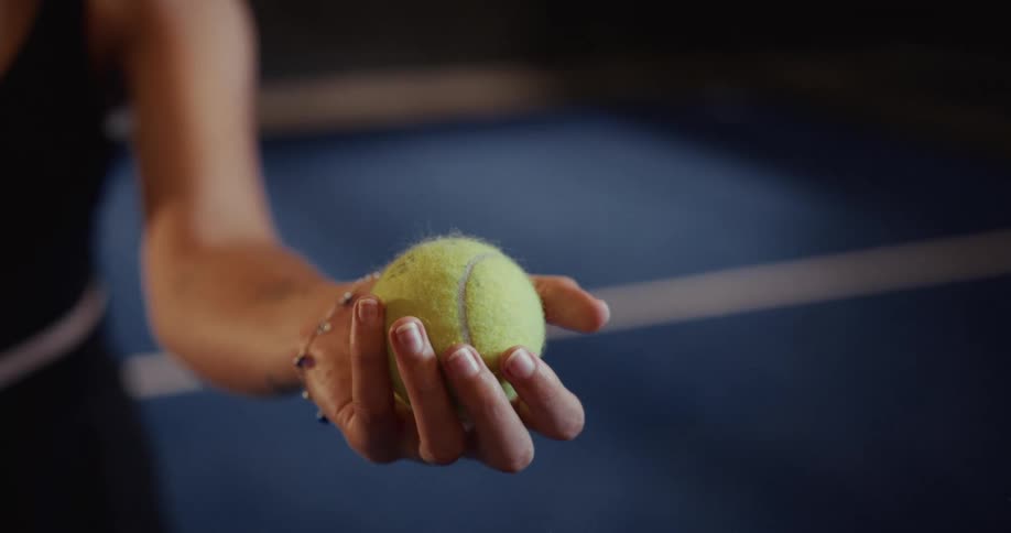 A female tennis player's hand holds a tennis ball steady on a blue court before a match