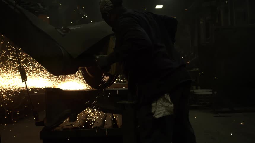 A factory worker in protective gear uses a large grinder, sending a cascade of sparks flying in slow motion in a dark workshop